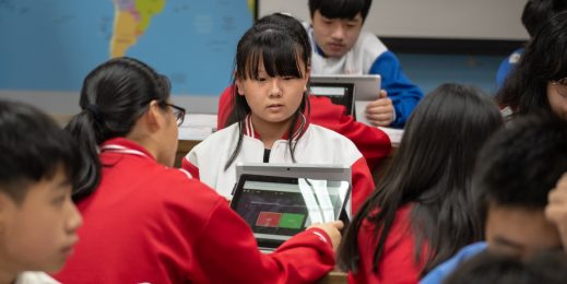 The camera focuses on a girl student in a classroom full of students.